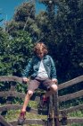 Menino da idade elementar com cabelo louro encaracolado sentado na ponte de madeira no campo . — Fotografia de Stock