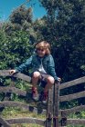 Menino da idade elementar com cabelo louro encaracolado sentado na ponte de madeira e sorrindo no campo . — Fotografia de Stock
