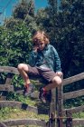Menino da idade elementar com cabelo louro encaracolado sentado na ponte de madeira no campo . — Fotografia de Stock