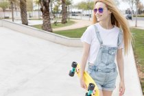Smiling teenage girl in sunglasses and denim overalls walking with penny board in summer park — Stock Photo