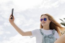 Ragazza bionda seduta e scattare selfie davanti al cielo nuvoloso — Foto stock