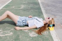 Blonde girl in sunglasses and jeans overalls lying on sidewalk with penny board under head — Stock Photo