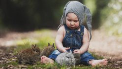 Cute little child in gray hat sitting with bunnies in park — Stock Photo
