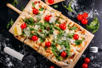 Rechteckig geschnittene Pizza mit Kirschtomaten, Kartoffeln und Käse auf Holzbrett auf dunklem Tisch — Stockfoto