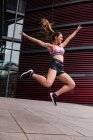 Aufgeregte, muskulös durchtrainierte Frau in Sportkleidung springt glücklich mit den Händen auseinander auf gepflasterter Straße — Stockfoto