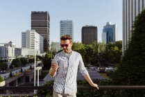 Eleganter Mann mit Sonnenbrille lehnt an Zaun und benutzt Smartphone in der modernen Stadt — Stockfoto