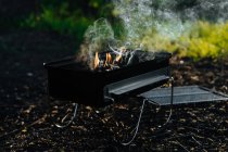 Grade de metal com chama ardente de carvão vegetal e fumaça colocada no chão em madeiras — Fotografia de Stock