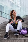 Fitte Frau in stylischer Sportbekleidung sitzt mit Tasche auf Stufen mit Apfel und Wasser — Stockfoto