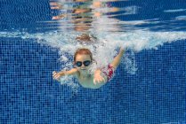 Дошкольник в очках плавает в голубом бассейне под водой с воздушными пузырьками — стоковое фото