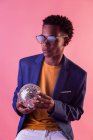 Black man wearing jacket and sunglasses holding glowing disco ball on pink background — Stock Photo