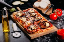 Pizza cortada con queso, salsa y berenjenas en rodajas sobre tabla de madera sobre mesa oscura - foto de stock
