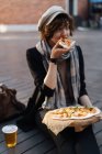 Молодая женщина сидит на подиуме на улице со стаканом пива и ест пиццу — стоковое фото
