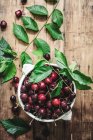 Flat lay of ceramic bowl filled with cherries and composed with green leaves on rustic table — Stock Photo