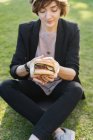 Молода жінка тримає бургер, сидячи на траві в парку — стокове фото