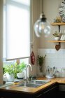 Light wooden kitchen interior with sink, stove and wooden cutting boards with basket and other dishware with potted plants on sill — Stock Photo