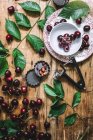 Puesta plana de cerezas, cherry pitter y hojas verdes sobre mesa rústica - foto de stock