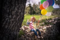 Vorschulkind sitzt mit bunten Luftballons auf Steinumrandung — Stockfoto