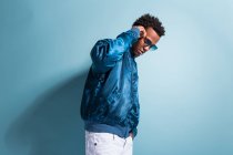 Cool black man in blue outfit and sunglasses standing on blue background — Stock Photo