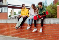 Веселі чоловічі та жіночі багатоетнічні підлітки сидять зі скейтбордами на цегляному паркані — стокове фото