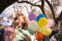 Preschooler boy with mouth open sitting on tree with balloons — Stock Photo