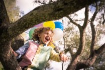 Bambino prescolare con la bocca aperta seduto su un albero con palloncini — Foto stock