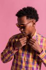 Young African American man in trendy multicolored checkered shirt and sunglasses standing on pink background — Stock Photo