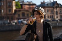 Вид сбоку стильной девушки в кепке, держащей пластиковый стаканчик с пивом и стоящей на городской набережной, отводящей взгляд от солнца — стоковое фото