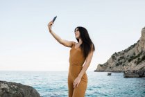Trendy brunette in fashionable overall taking selfie on cliff of seascape — Stock Photo