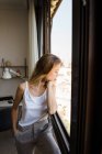 Вдумлива молода жінка стоїть і дивиться через вікно з головою на руці — стокове фото