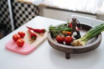 Свежие овощи и травы в деревянной миске и доске для рубки на кухонном столе — стоковое фото