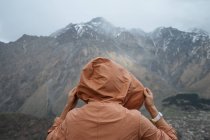 Back view of male in warm clothes with backpack hiking in mountains standing on grass looking at jagged mountain ridge covered with snow and peaks hidden in clouds — Stock Photo
