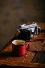 Enamel cup of coffee on rustic wooden surface with retro camera — Stock Photo