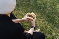 Mulher elegante sentado na grama do parque e segurando delicioso hambúrguer takeaway — Fotografia de Stock