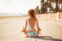 Visão traseira da mulher magra em maiô colorido e óculos de sol sentados na areia e olhando para longe — Fotografia de Stock