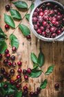 Flat lay of ceramic bowl filled with cherries and composed with green leaves on rustic table — Stock Photo