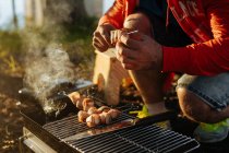 Чоловіки руками, підготовку бекон і ковбаси на рожні на грилі на спалювання вугілля в портативних сковорідка на відкритому повітрі — стокове фото