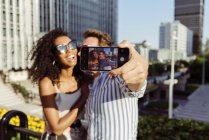 Alegre pareja multirracial posando para selfie mientras de pie en el fondo de la ciudad moderna - foto de stock