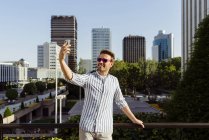 Hombre apoyado en la valla en la ciudad moderna y tomar selfie con teléfono inteligente - foto de stock