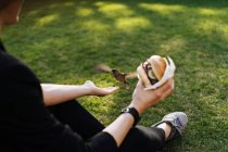 Woman sitting on green grass in park with takeaway burger and feeding sparrow — Stock Photo