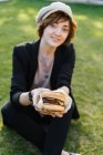 Lächelnde Frau zeigt Burger, während sie im Park auf Gras sitzt — Stockfoto