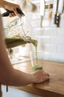Mani femminili versando sano frullato verde dalla tazza del frullatore in vetro sul bancone di legno in cucina — Foto stock