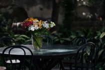 Elegant colorful assorted flowers in bouquet standing in glass vase with water on sunlit round black table with plants on blurred background - foto de stock
