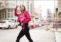 Mujer delgada en chaqueta de cuero rosa cruzando la calle y mirando hacia otro lado - foto de stock