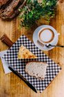 Café en taza de porcelana blanca en platillo con tabla de cortar a cuadros con rebanada de pan y trozo de pastelería - foto de stock