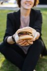 Sorrindo mulher mostrando hambúrguer enquanto sentado na grama no parque — Fotografia de Stock