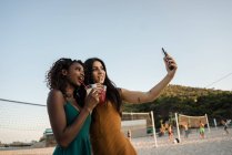 Young women taking selfie with drinks on sandy town beach — Stock Photo