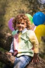 Sorridente bambino in età prescolare seduto su un albero con palloncini colorati — Foto stock