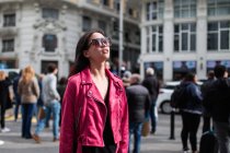 Trendy giovane donna moderna che indossa giacca di pelle rosa con occhiali da sole in piedi sulla strada alla luce del sole — Foto stock