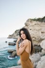 Side view of young stylish brunette in overall with long hair standing on cliffs of coastline and looking in camera — Stock Photo