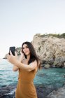 Stylish woman with long hair taking selfie on beach by sea water — Stock Photo